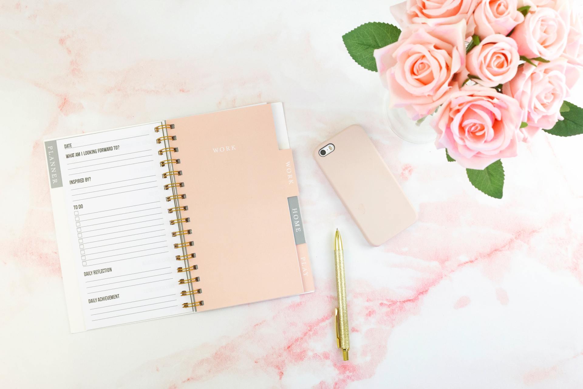A pink notebook , pen , cell phone and roses on a pink marble table.
