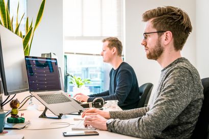 Two men are sitting at a desk in front of a laptop computer.