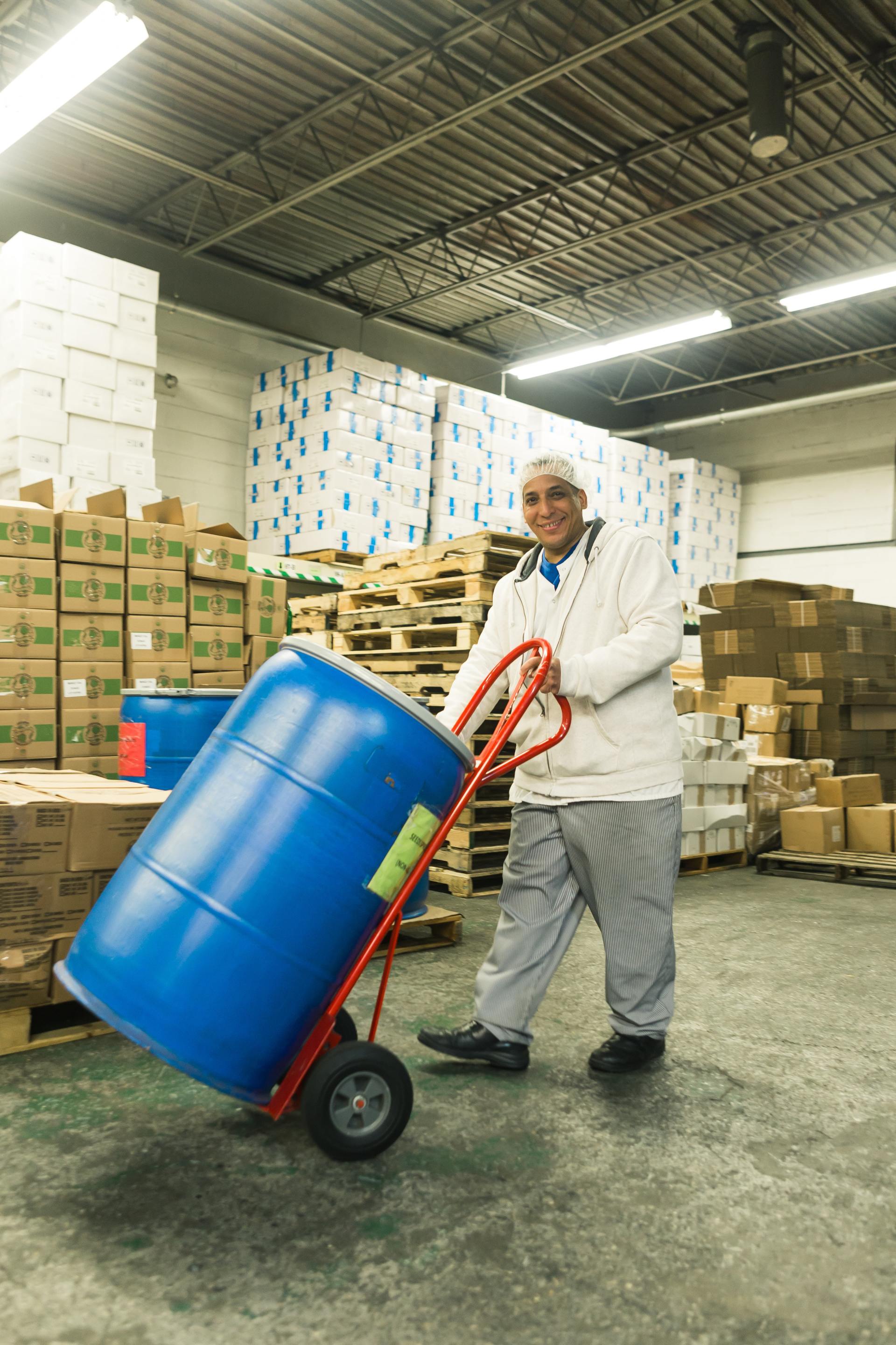 Man in work clothes smiles, pushing blue barrel on hand truck in a warehouse.