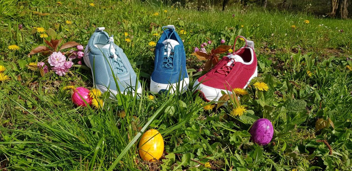 Drei bunte Turnschuhe halten Ostereier auf einer Wiese mit Löwenzahn.