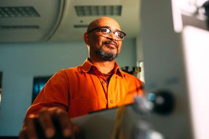 A man wearing glasses and an orange shirt is standing in front of a computer.