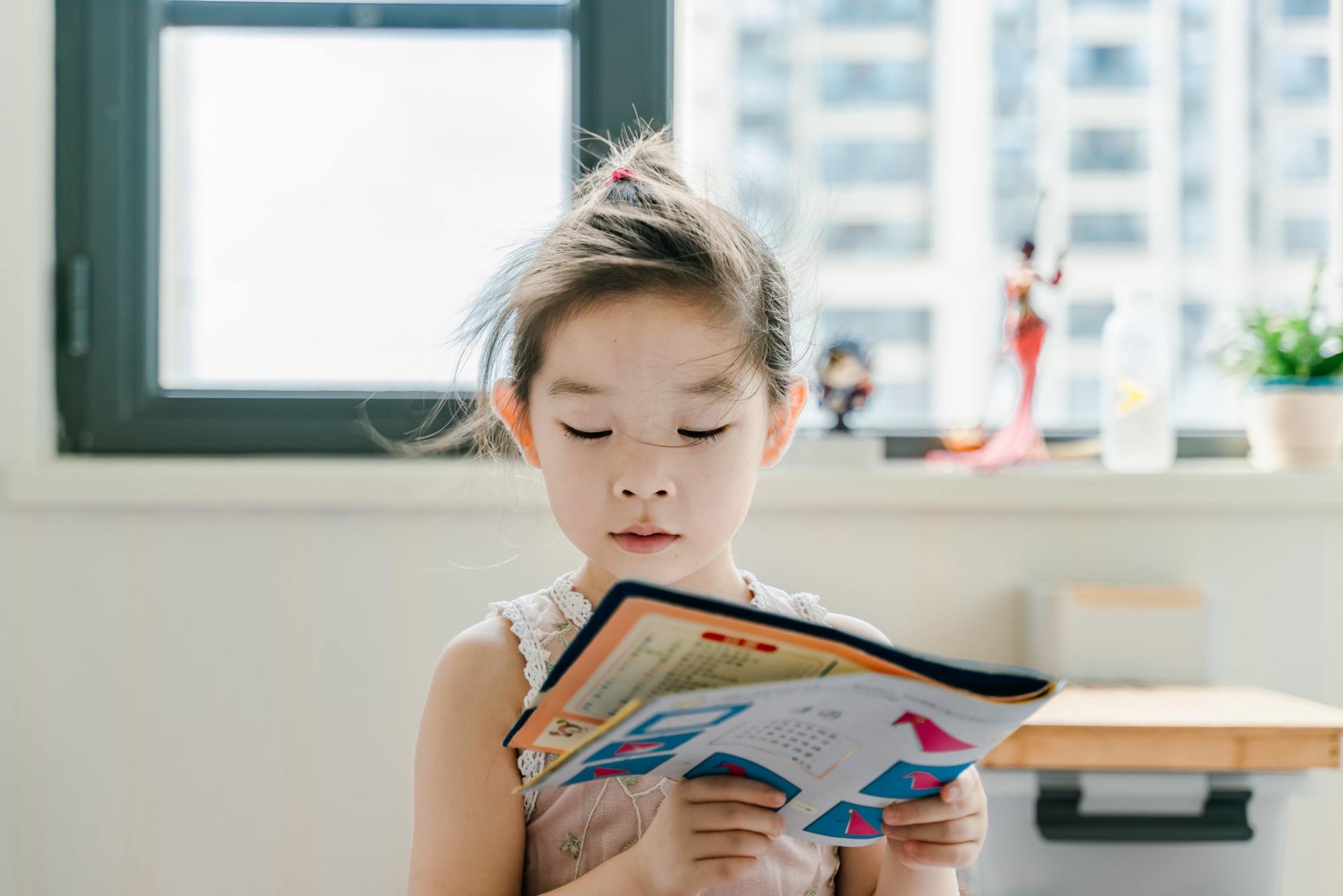 A little girl is reading a book in front of a window.
