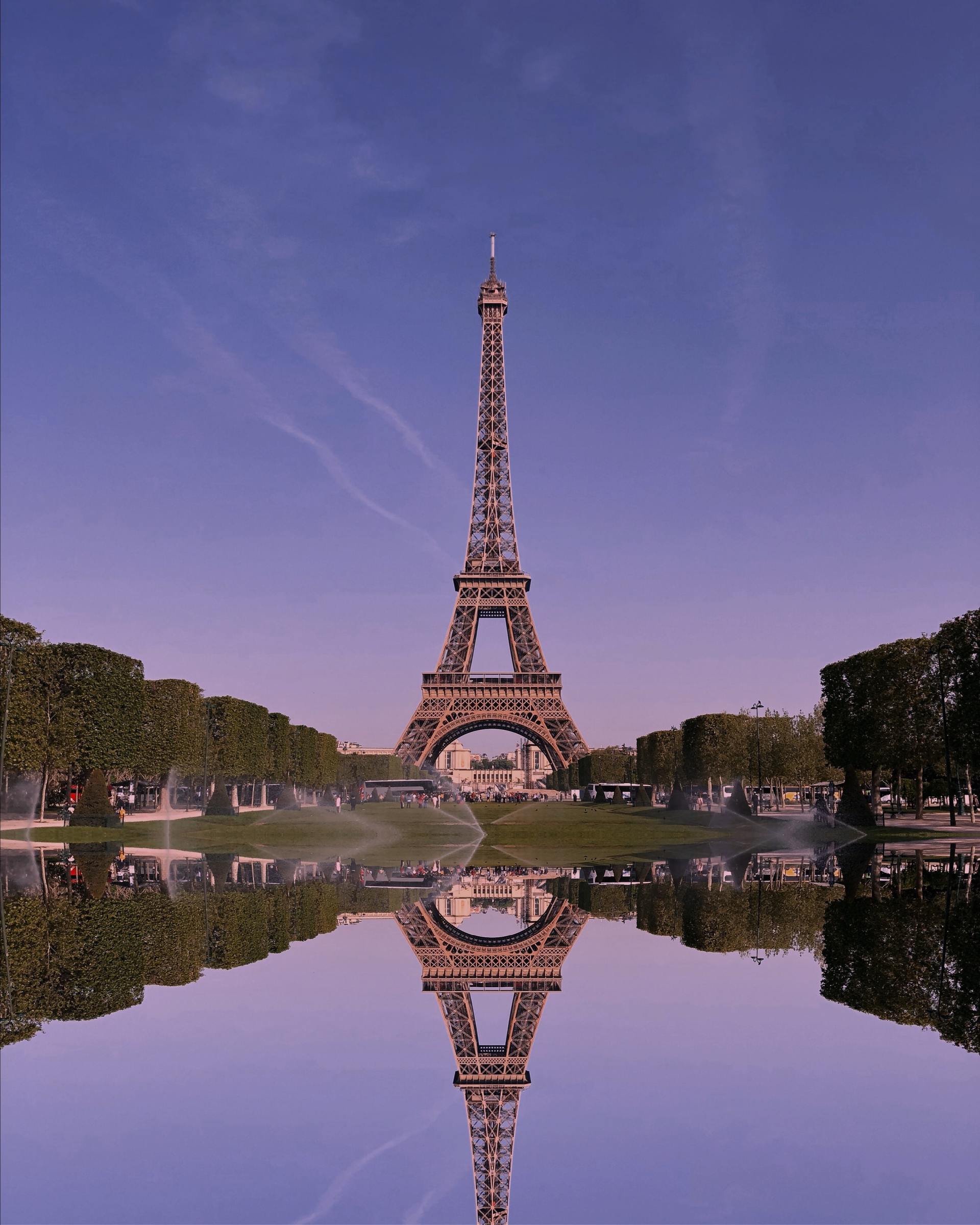 Eiffel Tower centered above a reflecting pool at sunset, mirrored in calm water with trees lining both sides