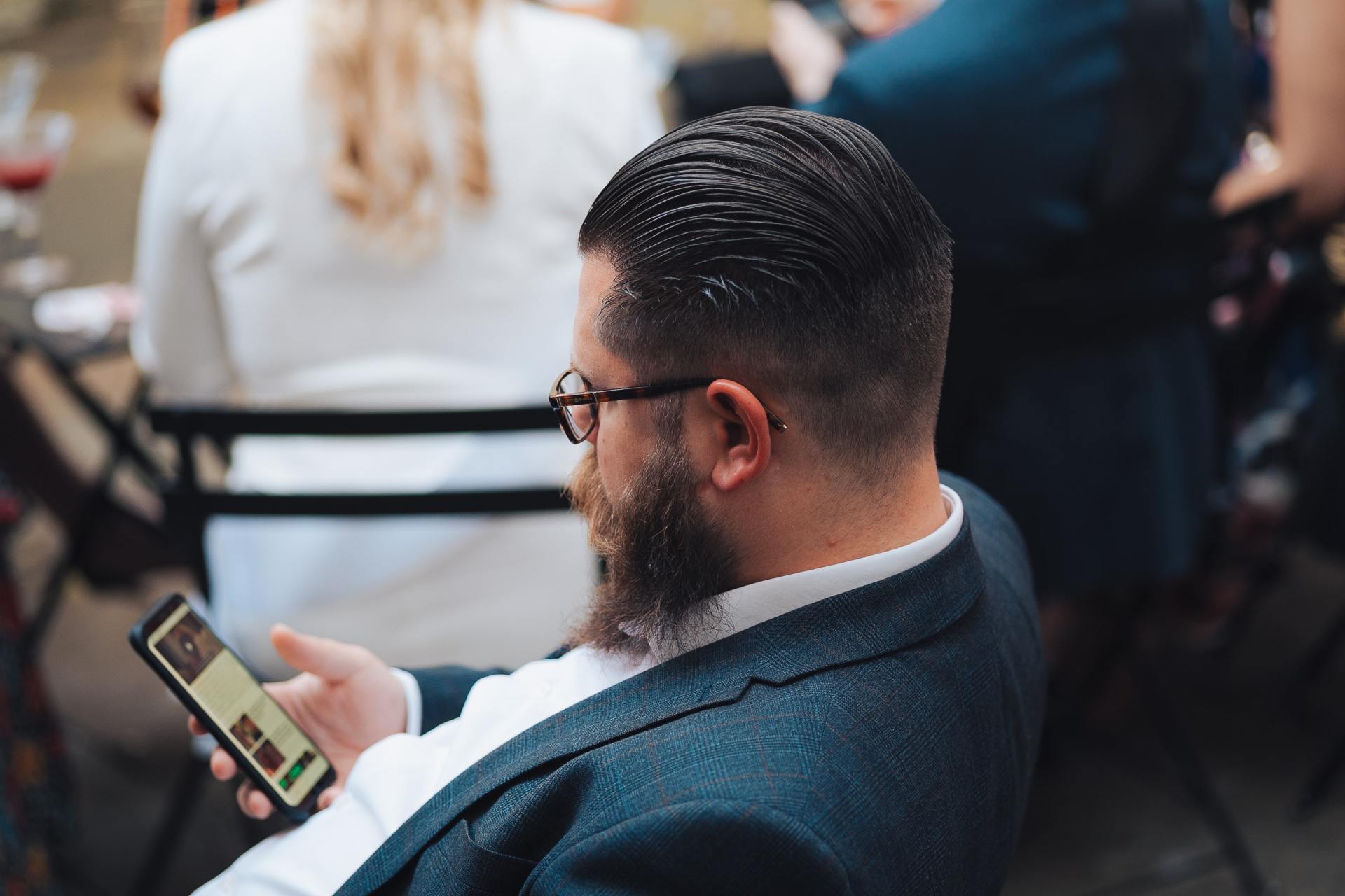 A man with a beard is sitting in a chair looking at his cell phone.
