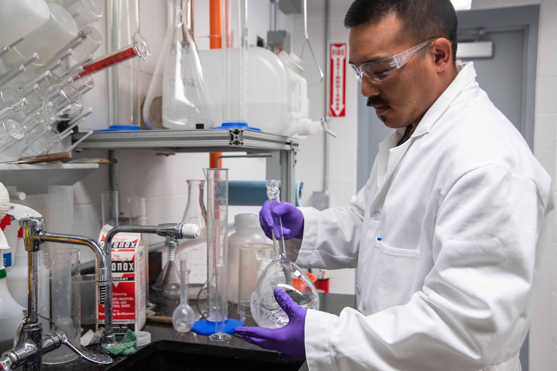 A man in a lab coat and purple gloves is pouring liquid into a beaker.