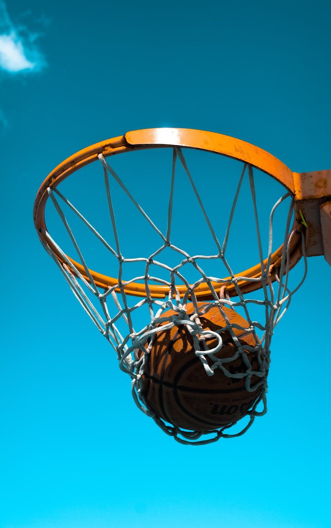 Basketball hoop with orange rim and net, viewed from below against a bright blue sky.