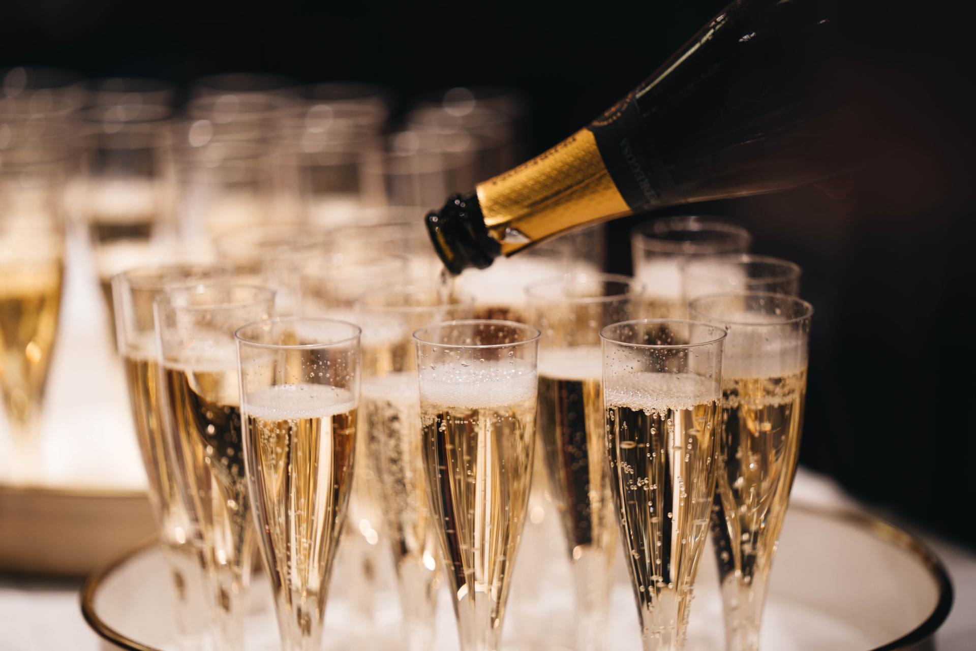 A bottle of champagne is being poured into a row of champagne flutes.