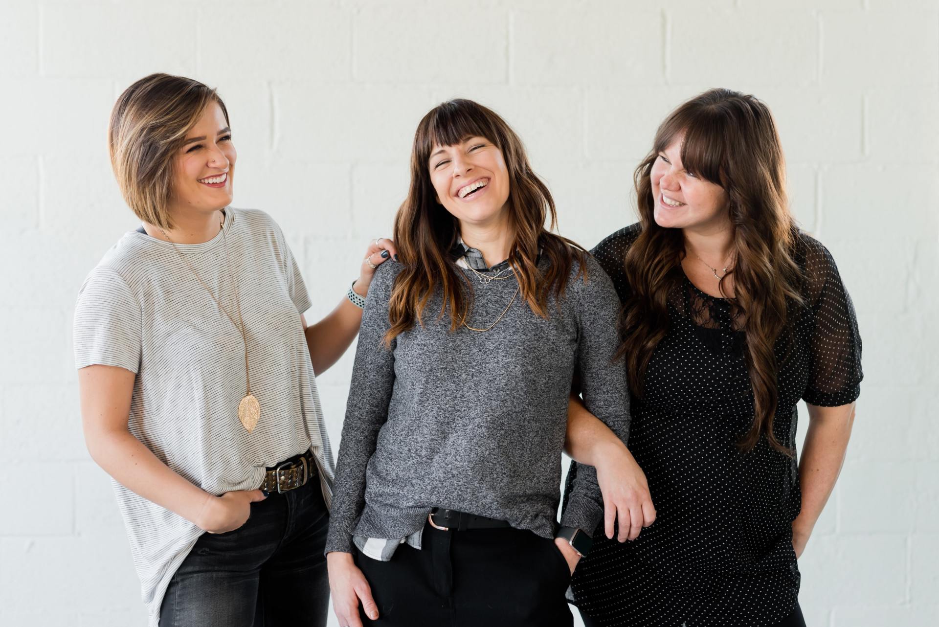 Three smiling women pose together in front of a white wall. 