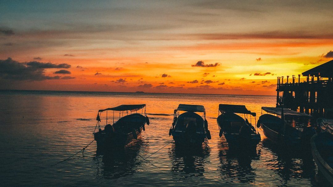 Boats silhouetted on water at sunset. Orange and yellow sky, calm sea.