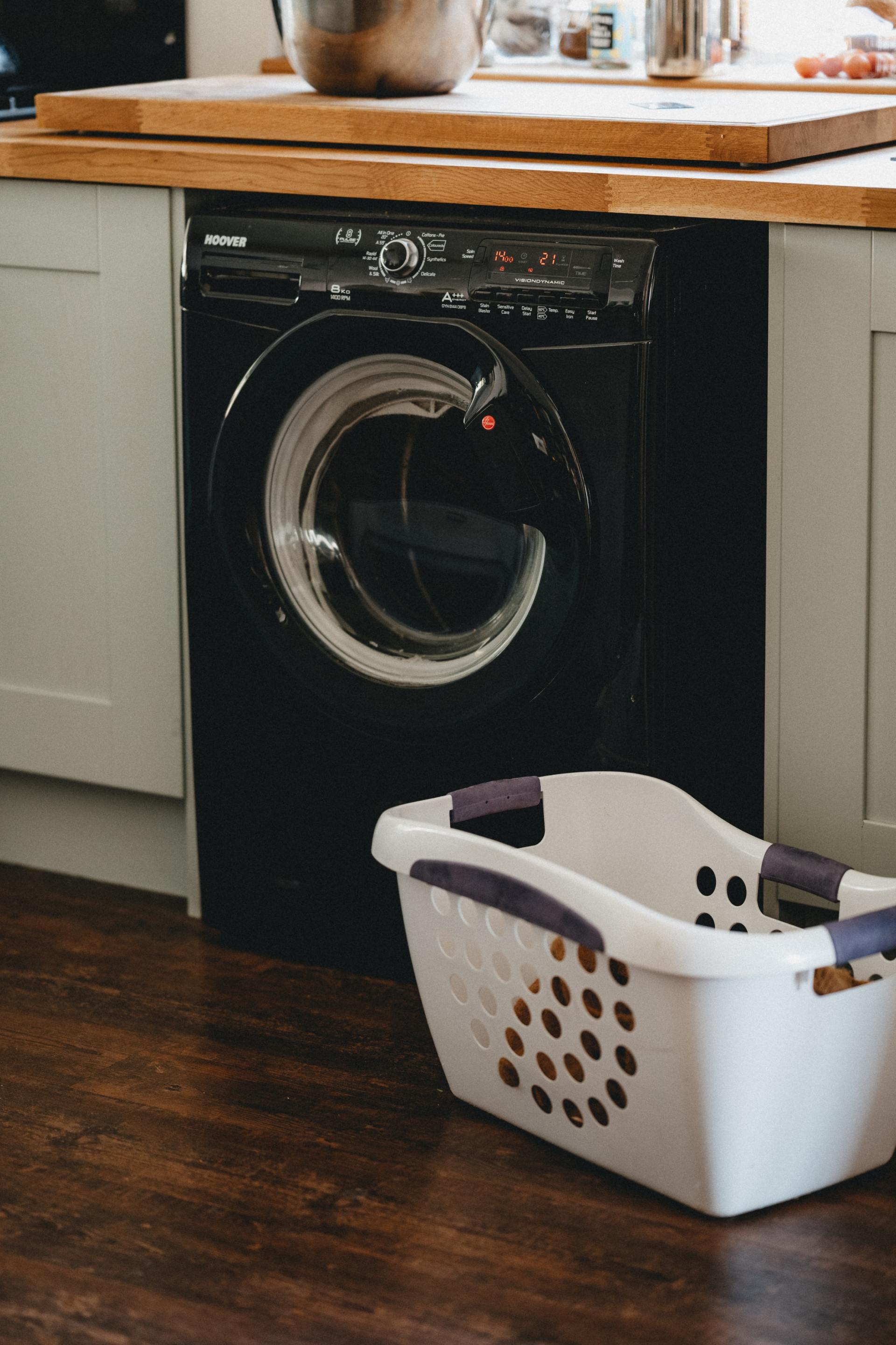 A black washing machine installed under a wooden kitchen countertop with a white laundry basket on the floor in front.
