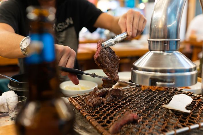 A person grilling meat with tongs at a table. A bottle of beer is in the foreground.