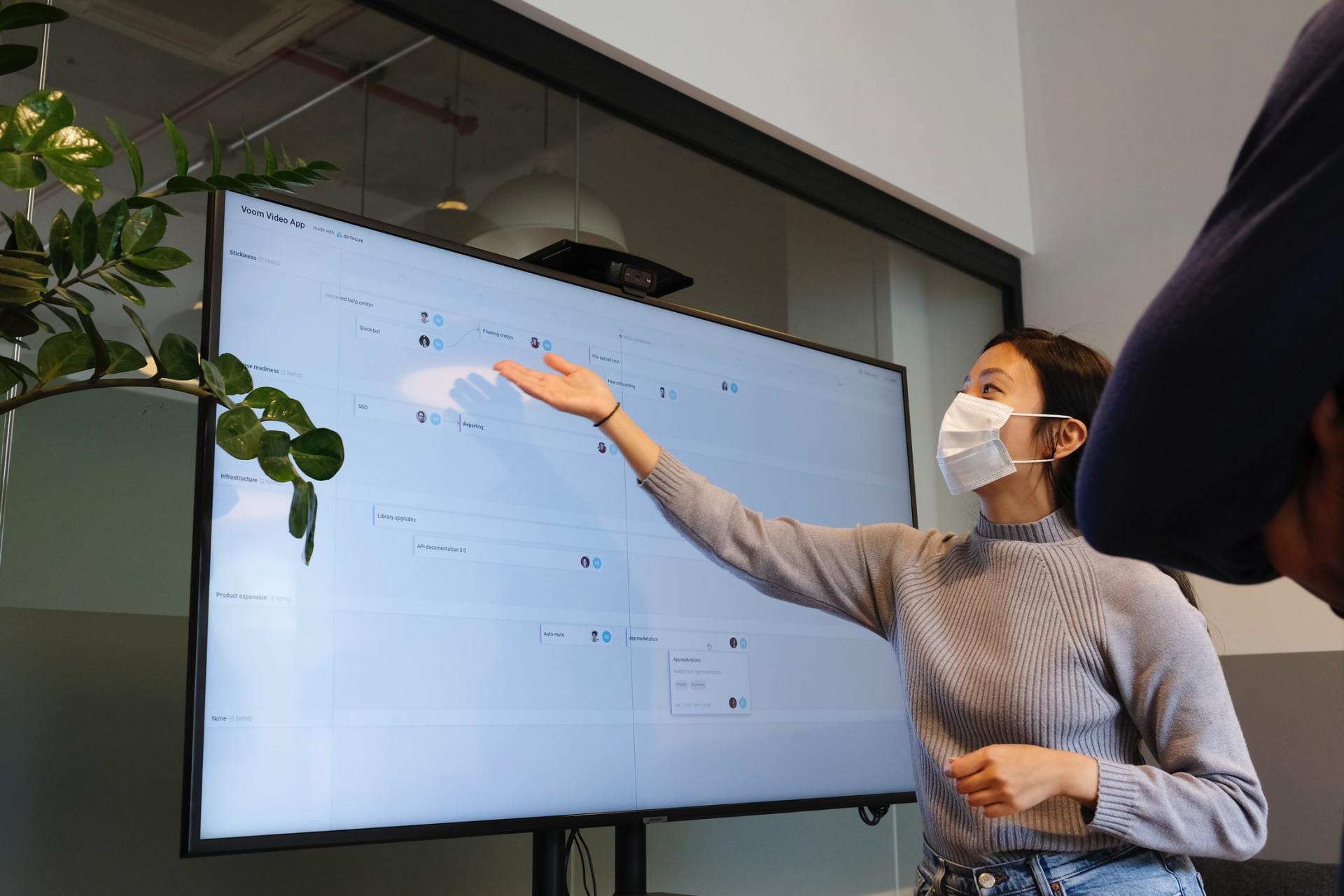 A woman wearing a mask is giving a presentation in front of a large screen.