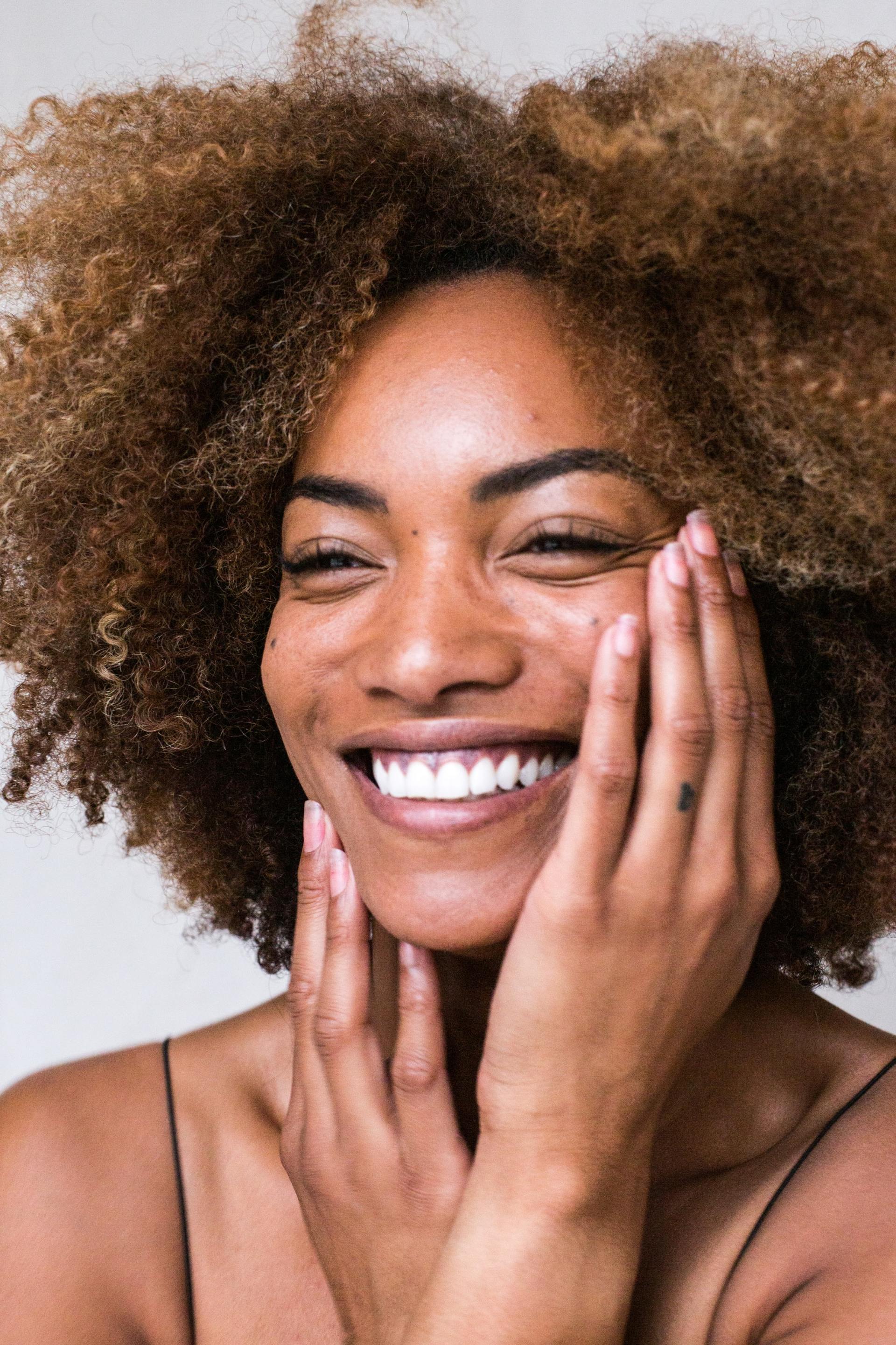 A woman with curly hair is smiling and touching her face.