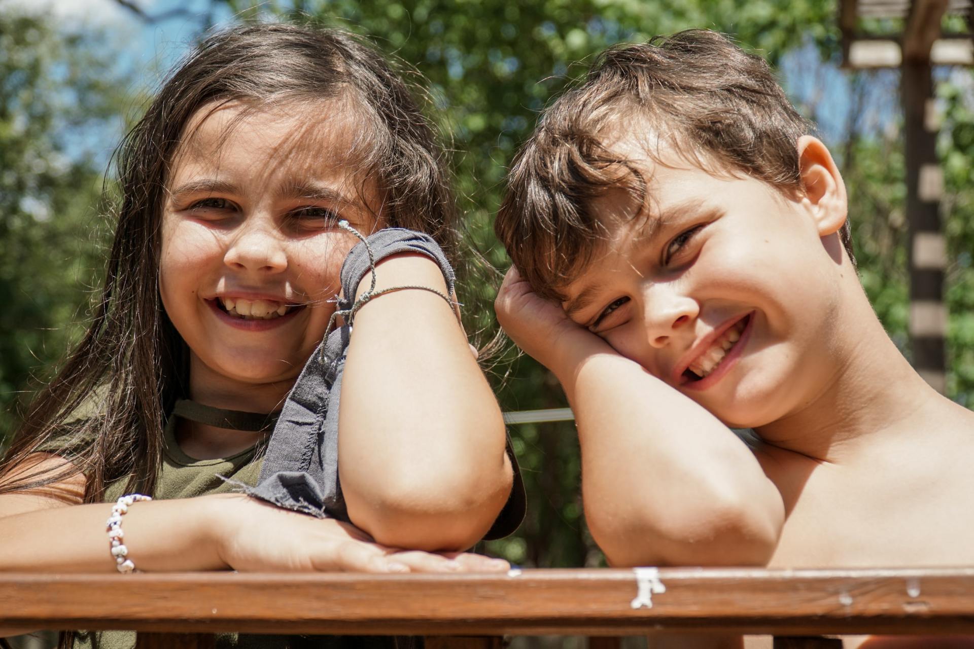 Two smiling children resting their heads on their hands outdoors behind a wooden fence