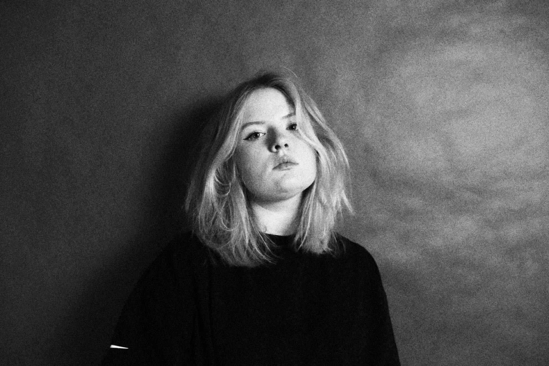 Woman with blonde hair, dark shirt, leaning against a textured gray wall, looking towards the camera.