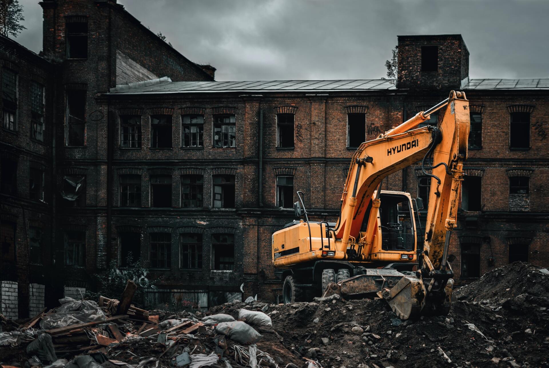 Yellow excavator at a decaying brick building demolition site under a cloudy sky.