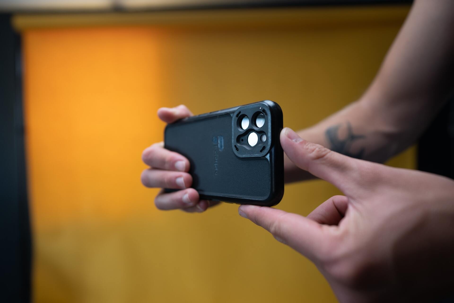 Person holding a black phone with a triple-lens camera, finger pointing to the lenses, against an orange background.