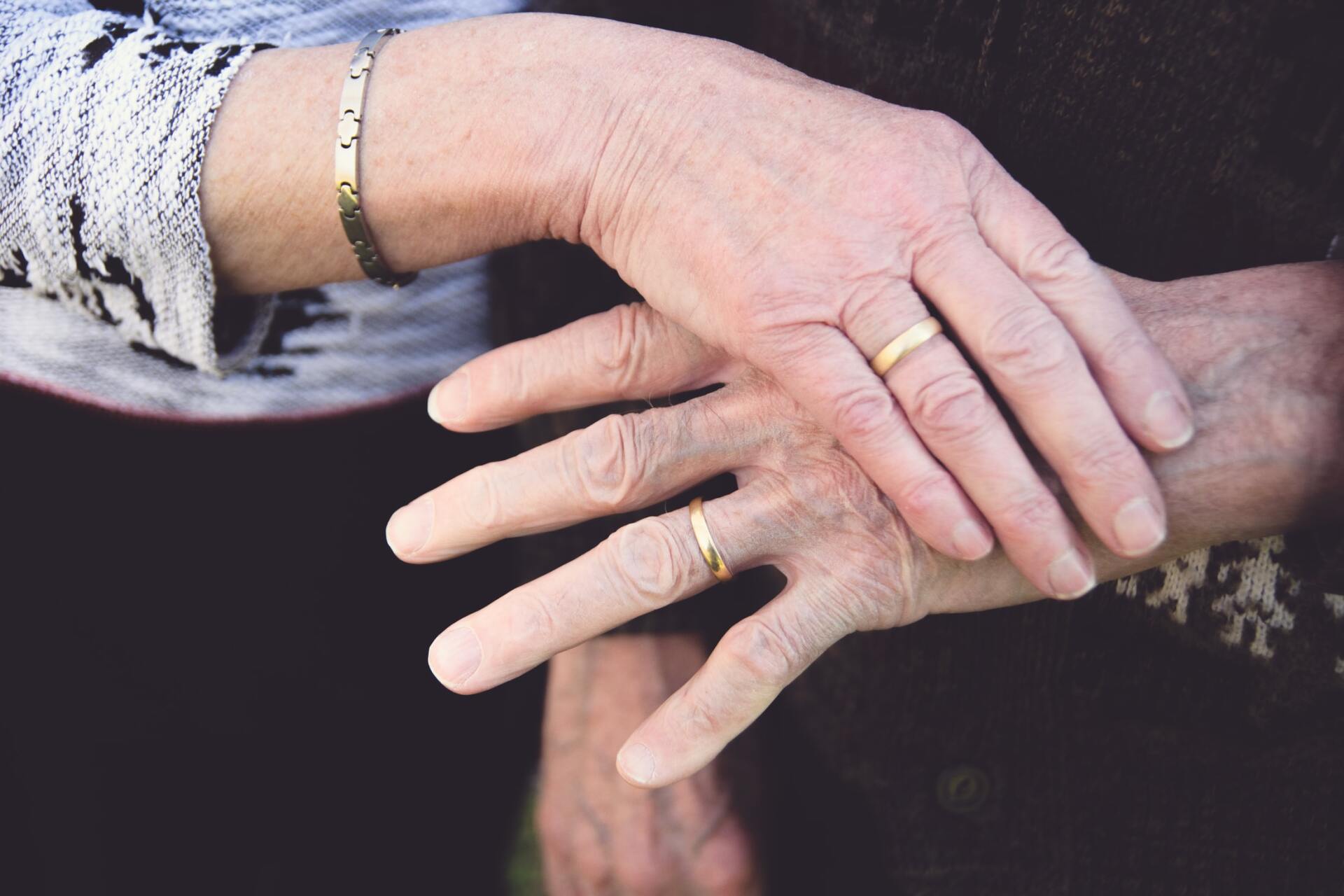 Two elderly hands gently clasped, with gold rings and a bracelet on a dark background.