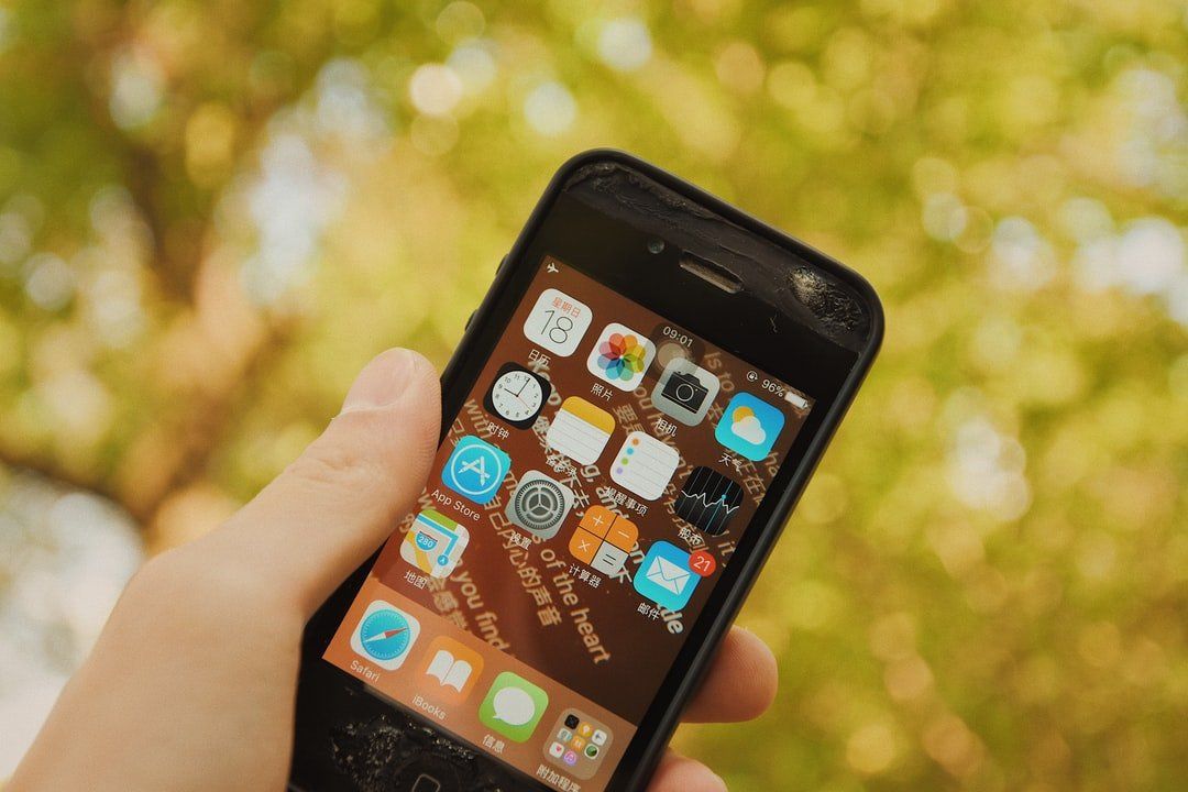 Hand holding a smartphone with app icons on the home screen outdoors, against a blurred leafy background