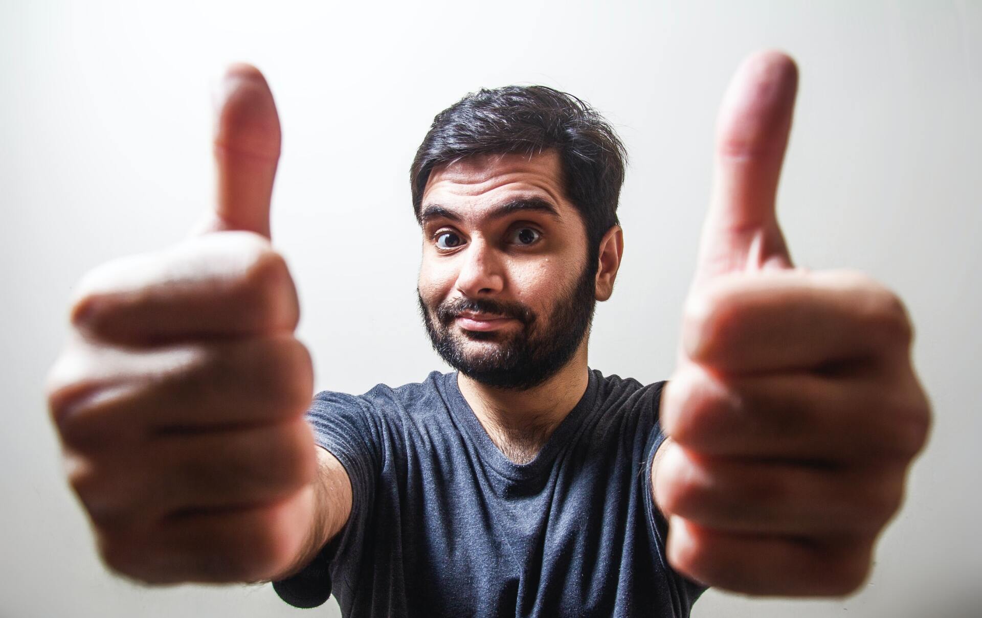 Man with dark hair and beard, wearing a gray t-shirt, giving two thumbs up.