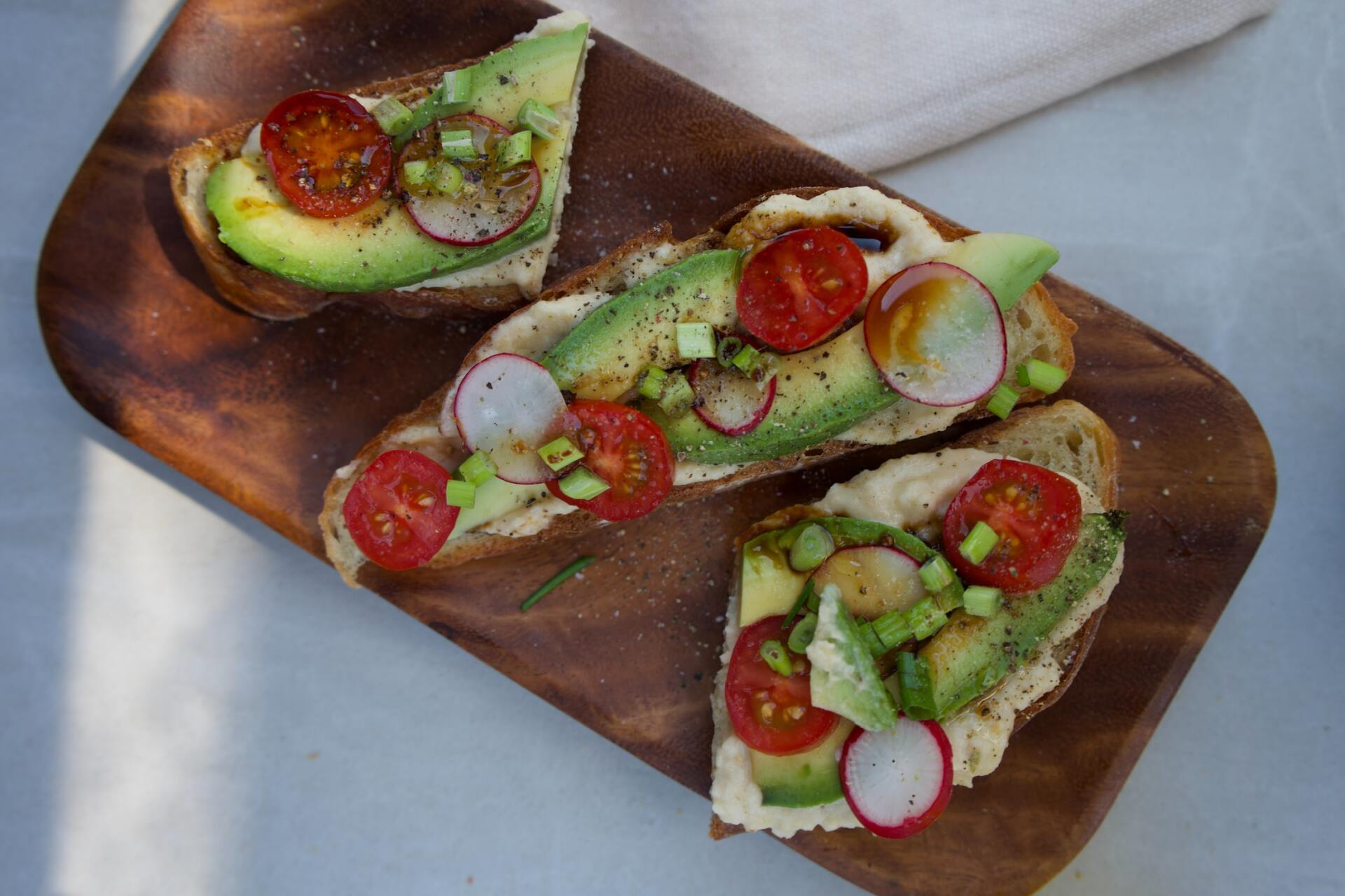 Three slices of toast with avocado , tomatoes and radishes on a wooden cutting board.