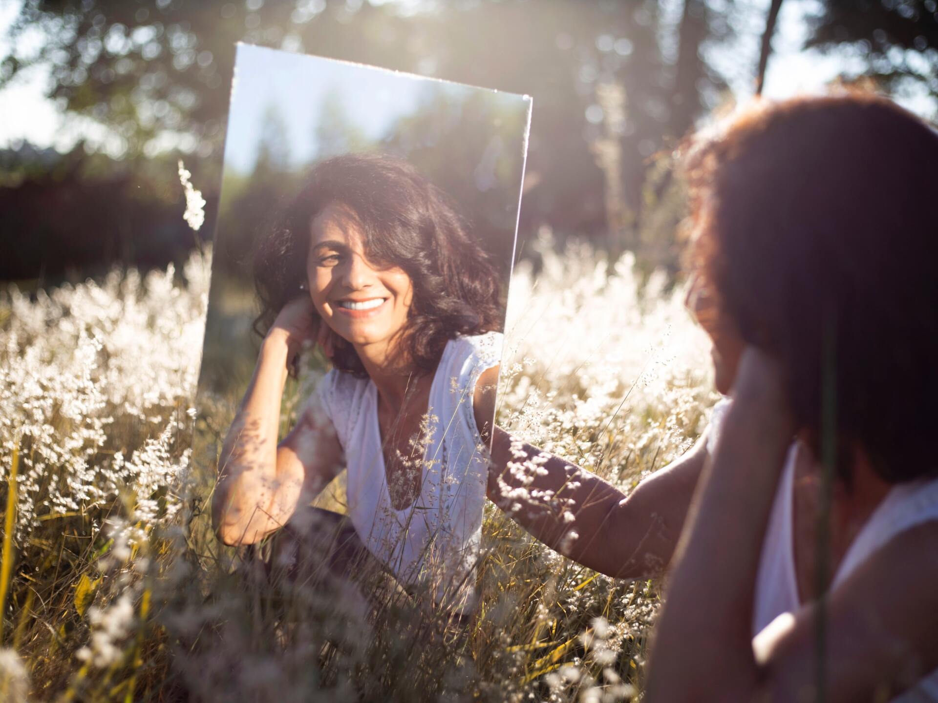Woman smiling in a sunlit field, reflected in a mirror held by another person