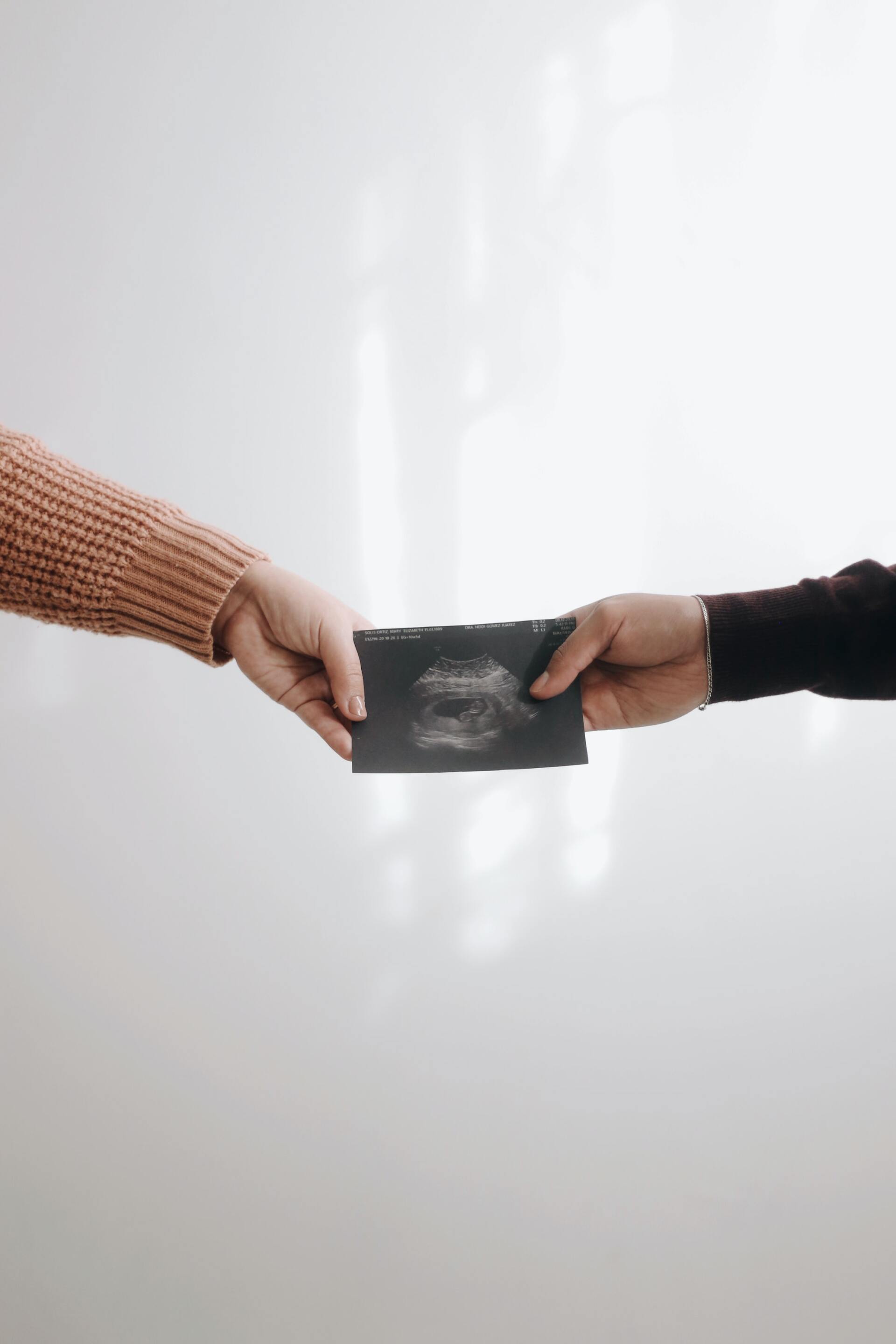 Hands passing a dark wallet against a white background. One arm wears a tan sweater, the other a black one.