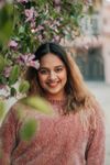 Woman smiles, standing in front of pink flowers, wearing a pink sweater.