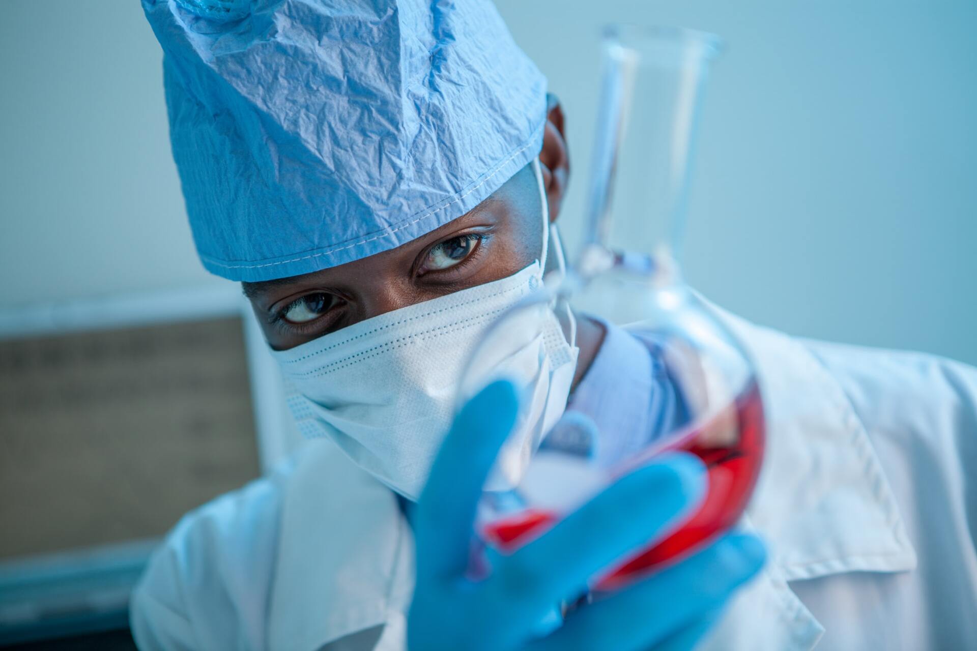 Scientist in lab coat, cap, mask, and gloves examining a flask filled with red liquid.
