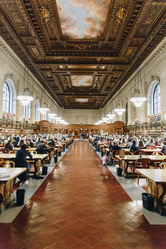 Grand reading room with long tables, people studying, ornate ceiling, and large arched windows