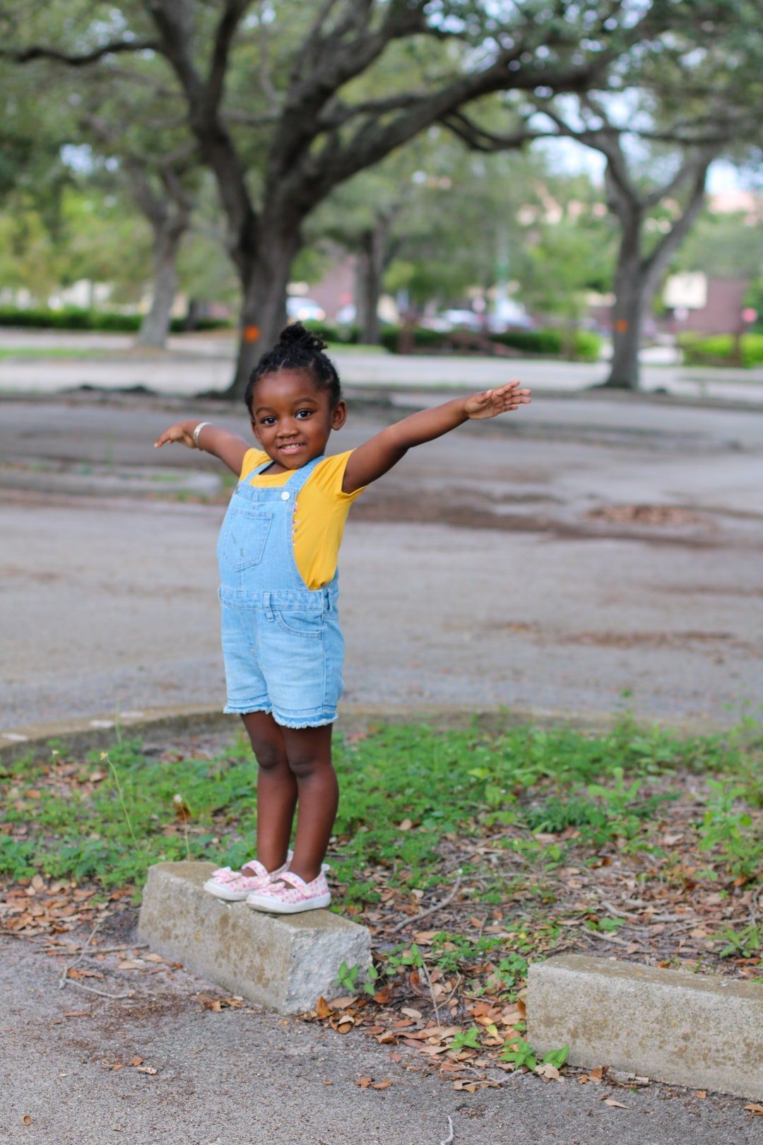 Young child with arms outstretched, standing on a curb in a park, wearing blue overalls and a yellow shirt.