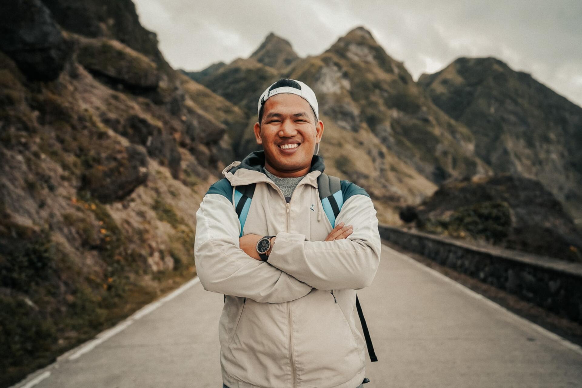 A man is standing on a road with his arms crossed in front of a mountain.