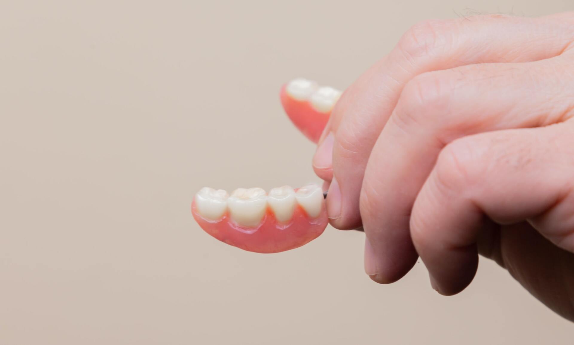 Hand holding a set of dentures, with pink gum-like base and white teeth, against a blurred beige background.