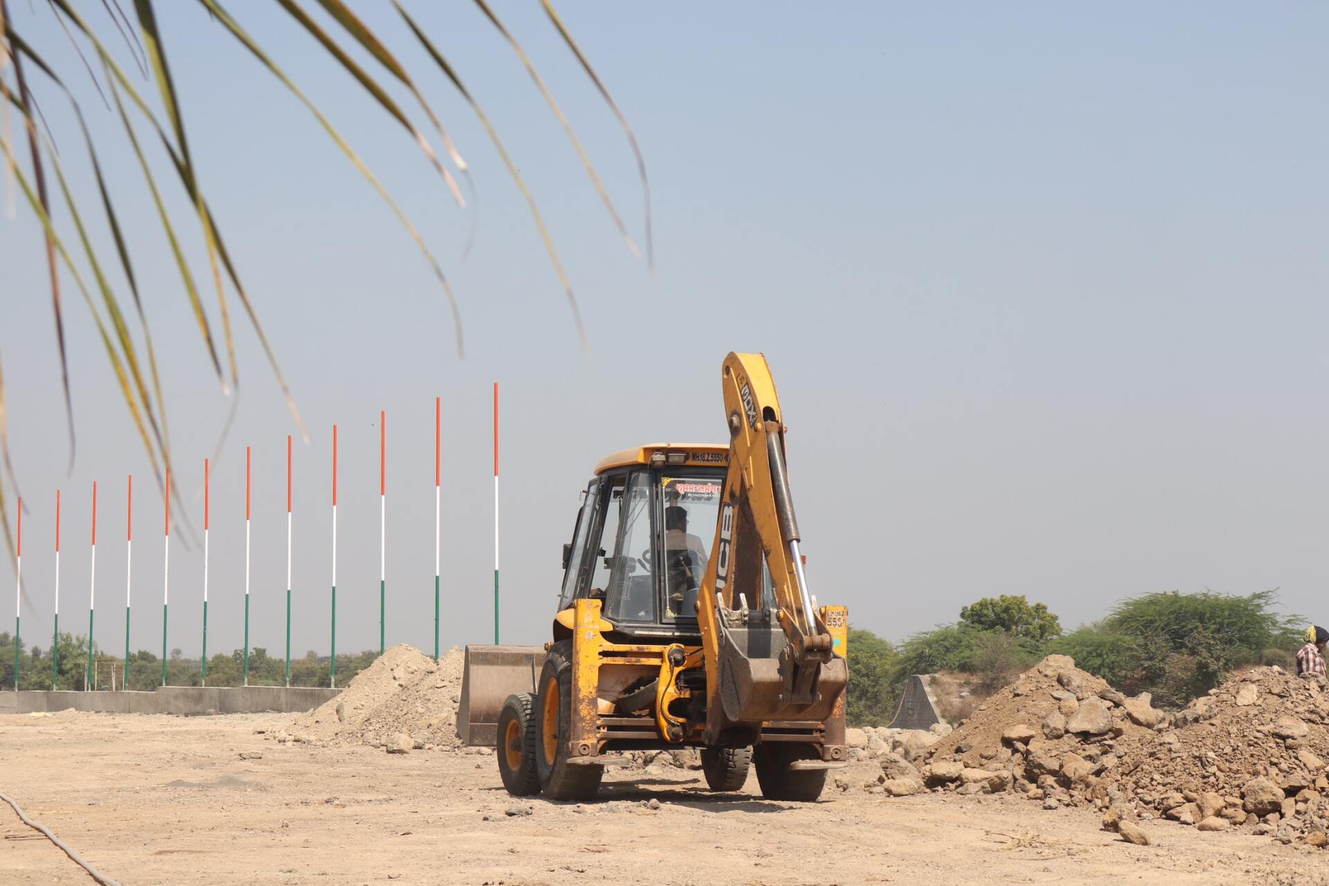 Yellow backhoe working on a construction site; dirt mounds in foreground, poles in background.