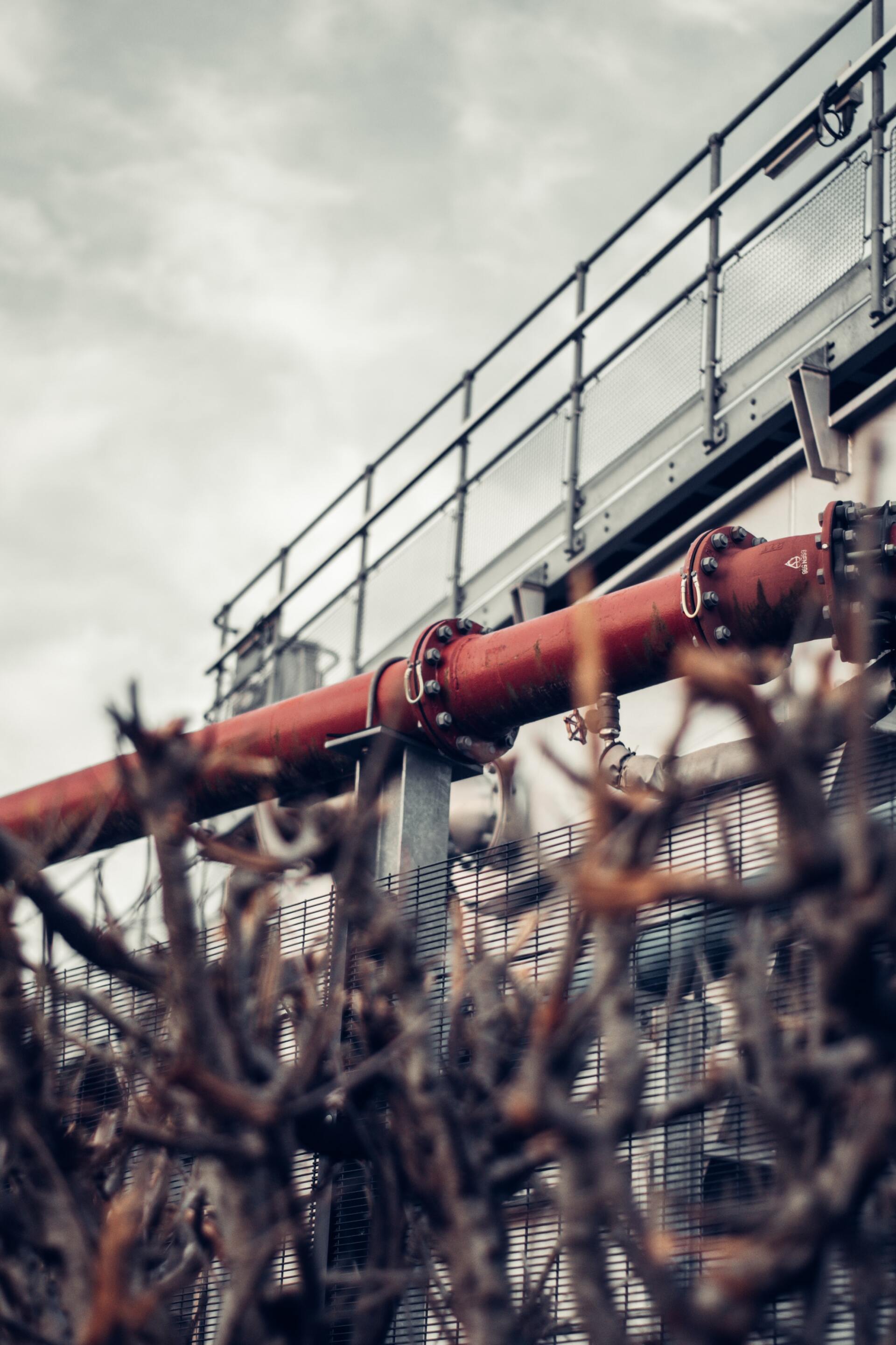 Red pipe runs along a building with a metal fence. Barbed wire in the foreground under a cloudy sky.
