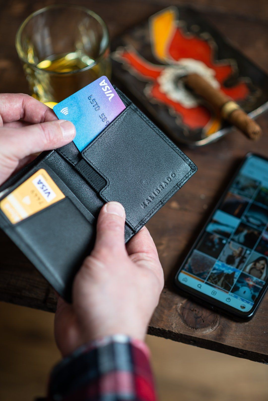 Person holding wallet with credit cards, a cigar and a phone on a wood table.