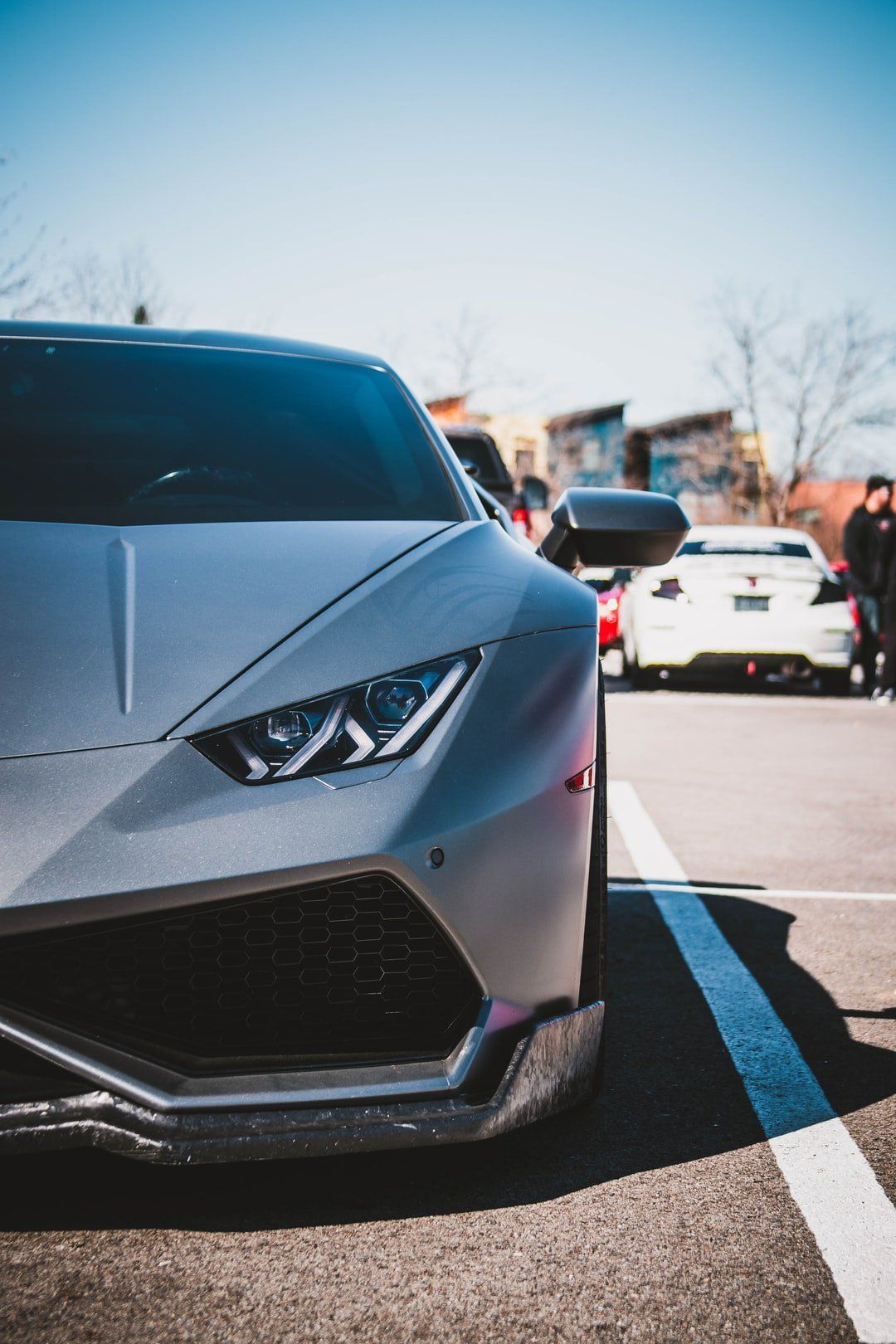 Silver sports car parked in a lot, with a white car and people blurred in the background