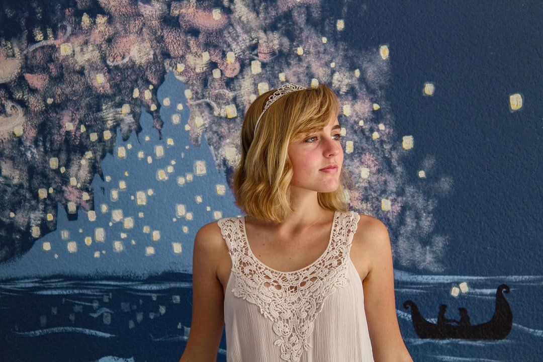 A woman in a white tank top is standing in front of a painting of a boat.