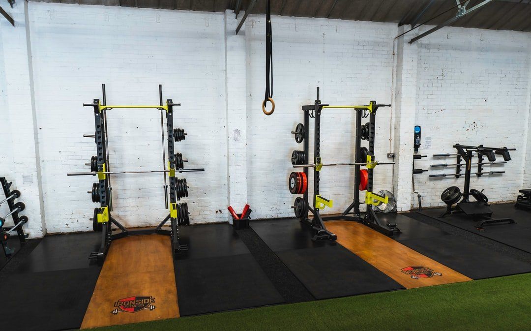 Gym with squat racks, barbells, mats, and hanging rings against a white wall